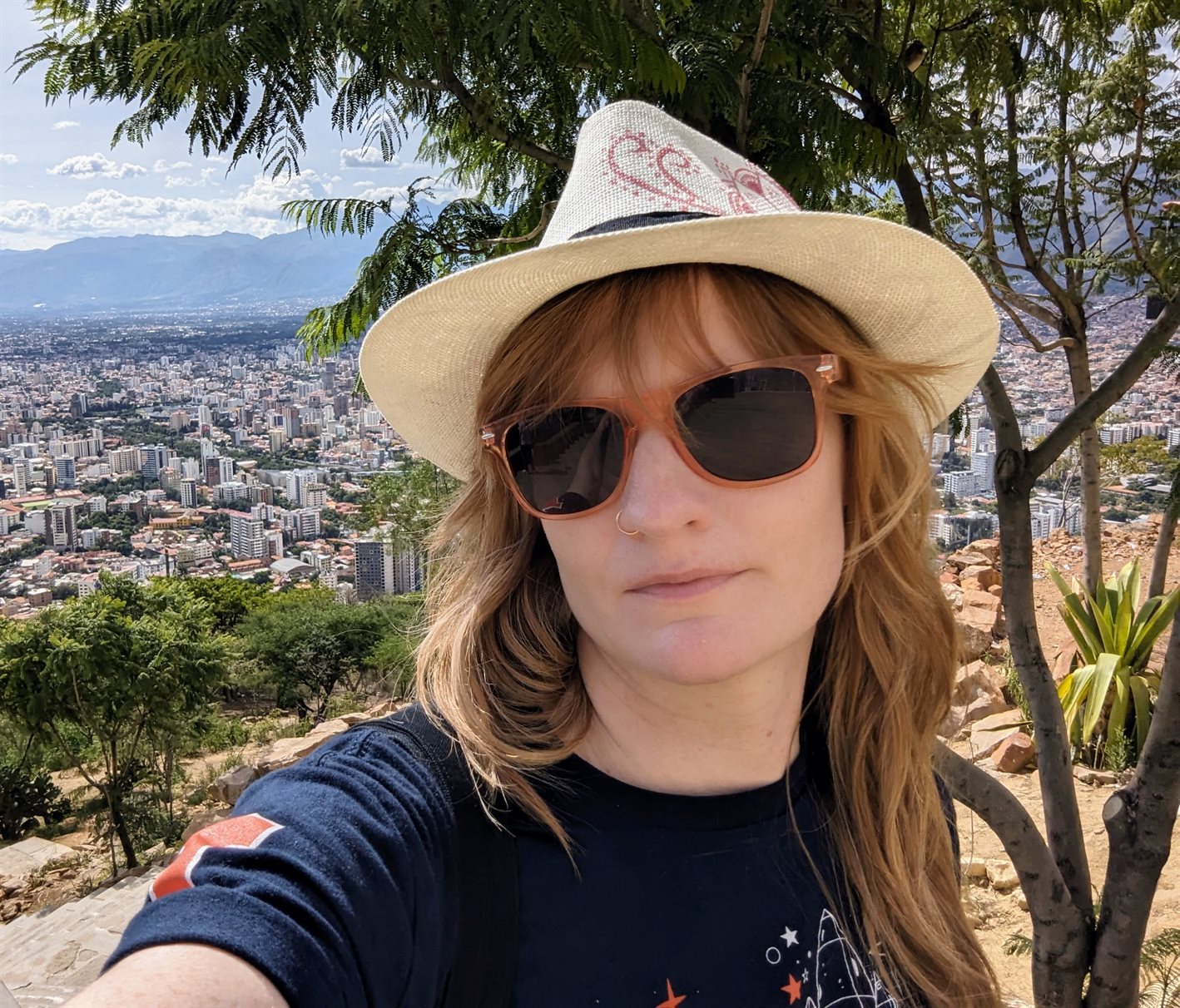 A selfie of pale white woman with red hair wearing a white hat, orange sunglasses, and a blue t-shirt in front of an overlook of a city.